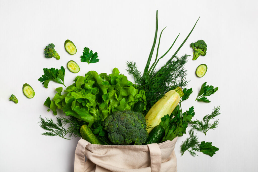 Un tote bage beige, d'où sortent différents légumes verts: salade, courgette, fenouille, céléri, brocoli et concombre.