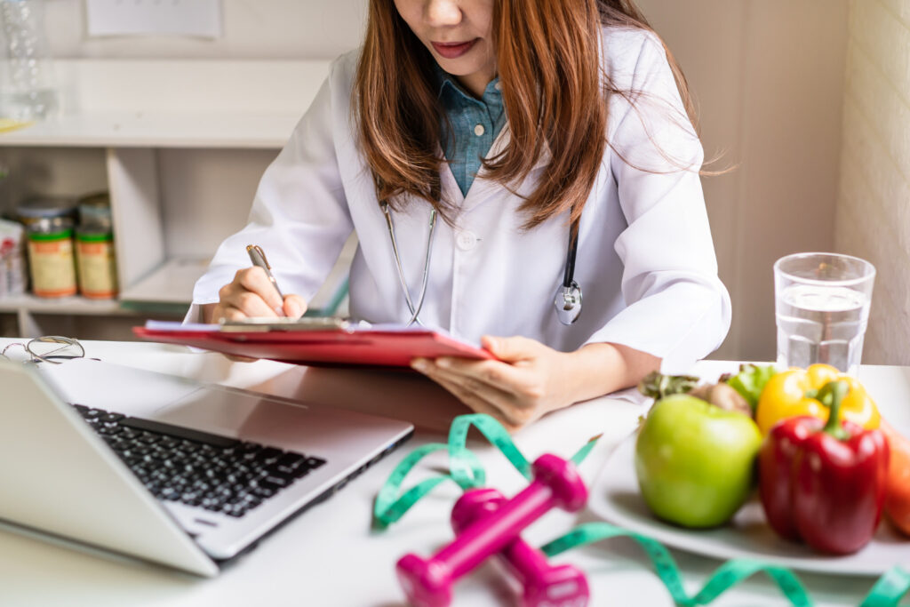 Un expert en nutrition entrain d'écrire sur un document devant un ordinateur portable. A côté d'elle se trouvent des fruits, un mètre ruban vert et des petits haltères de couleur magenta.