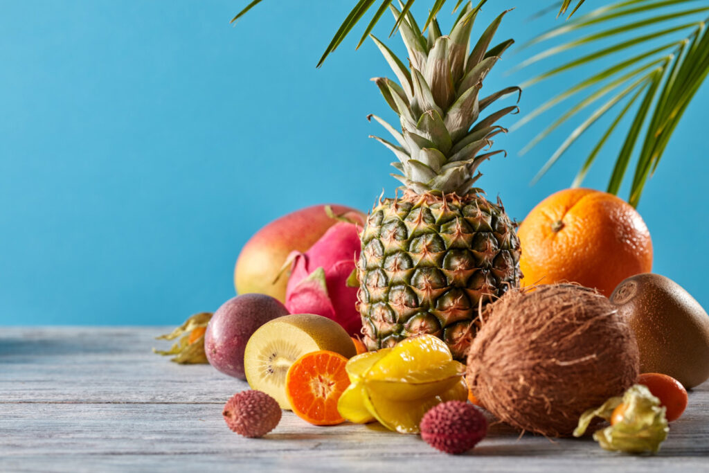 Divers fruits tropicaux (ananas, kiwi, coco, orange, mangue, litchis...) posés sur un support en bois, sur un fond bleu avec une feuille de palmier.